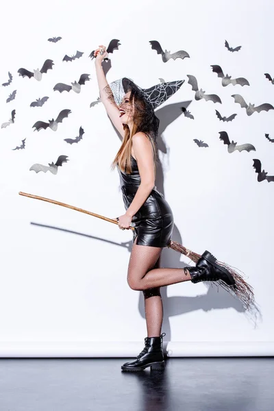 Счастливая девушка в костюме черной ведьмы Хэллоуин на метле возле белой стены с декоративными летучими мышами — стоковое фото