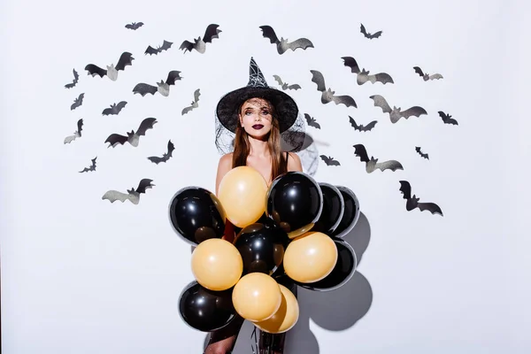 Girl in black witch Halloween costume holding balloons near white wall with decorative bats — Stock Photo