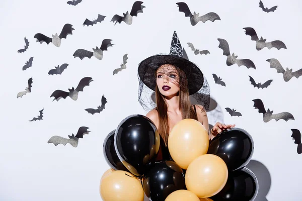 Mädchen in schwarzem Hexenhalloween-Kostüm mit Luftballons nahe weißer Wand mit dekorativen Fledermäusen — Stockfoto