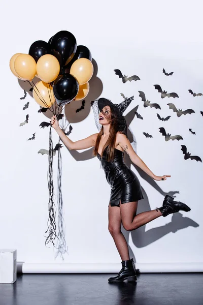 Menina em preto bruxa Halloween traje segurando balões perto da parede branca com morcegos decorativos — Fotografia de Stock