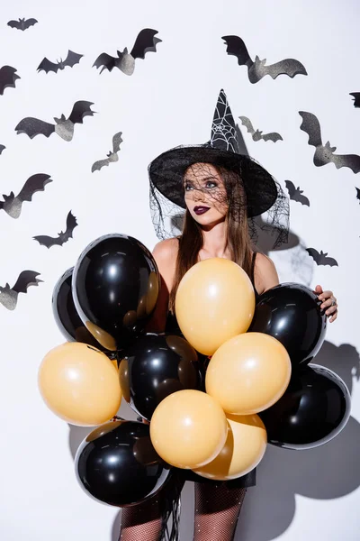 Mädchen in schwarzem Hexenhalloween-Kostüm mit Luftballons nahe weißer Wand mit dekorativen Fledermäusen — Stockfoto