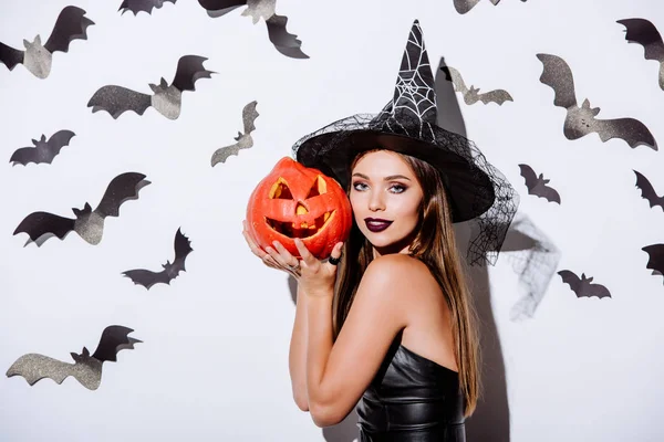 Girl in black witch Halloween costume holding carved spooky pumpkin near white wall with decorative bats — Stock Photo