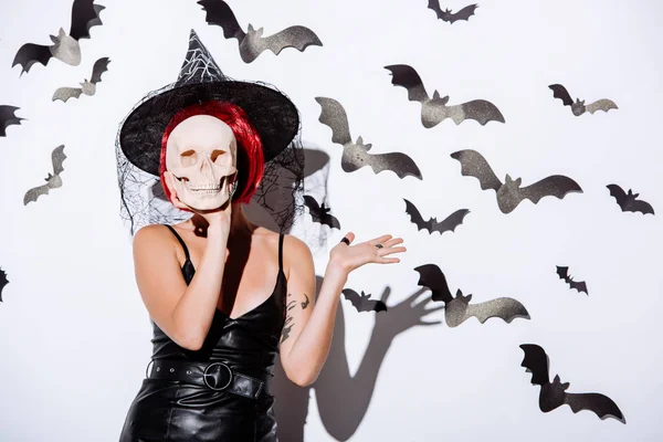 Chica en traje de Halloween bruja negro con el pelo rojo celebración de cráneo en frente de la cara cerca de la pared blanca con murciélagos decorativos - foto de stock