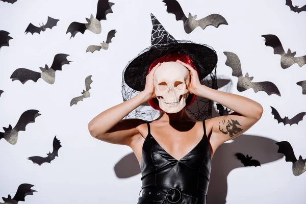 Girl in black witch Halloween costume with red hair holding skull in front of face near white wall with decorative bats — Stock Photo