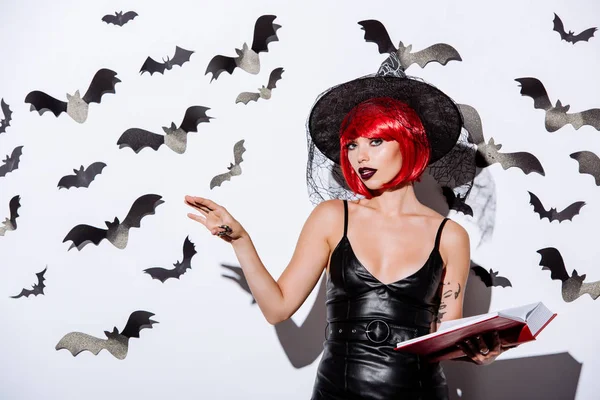 Chica en traje de Halloween bruja negro con pelo rojo celebración libro cerca de la pared blanca con murciélagos decorativos - foto de stock