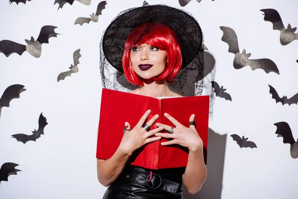 Girl in black witch Halloween costume with red hair holding book near white wall with decorative bats — Stock Photo