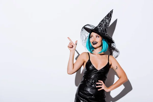 Chica sonriente en traje de Halloween bruja negro con el pelo azul señalando con el dedo sobre fondo blanco - foto de stock