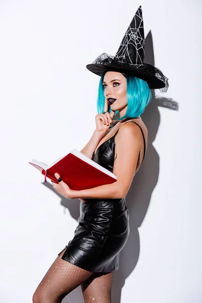 Сексуальна дівчина в чорній відьмі костюм Хеллоуїна з блакитним волоссям тримає книгу на білому тлі — стокове фото