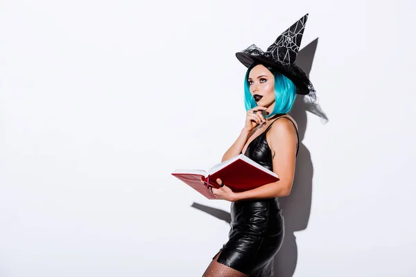 Ragazza sexy pensosa in costume strega nera Halloween con capelli blu libro tenuta su sfondo bianco — Foto stock