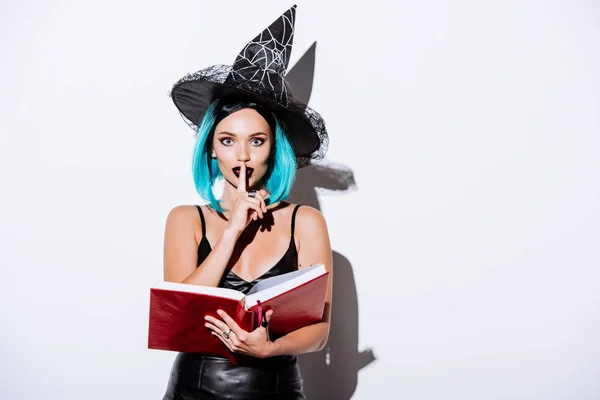 Sexy chica en negro bruja de Halloween traje con azul pelo celebración libro y mostrando shh gesto sobre fondo blanco - foto de stock