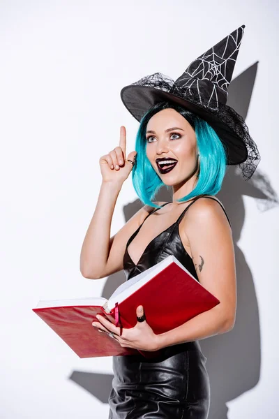 Збуджена сексуальна дівчина в чорному костюмі на Хеллоуїн з блакитним волоссям тримає книгу і показує жест ідеї на білому тлі — стокове фото