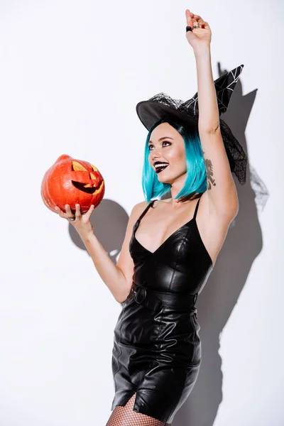 Fille sexy en noir sorcière Halloween costume avec des cheveux bleus tenant fantasmagorique citrouille sculptée sur fond blanc — Photo de stock