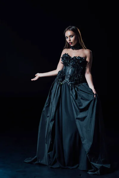 Asustadiza chica vampiro en vestido gótico negro apuntando con la mano sobre fondo negro - foto de stock