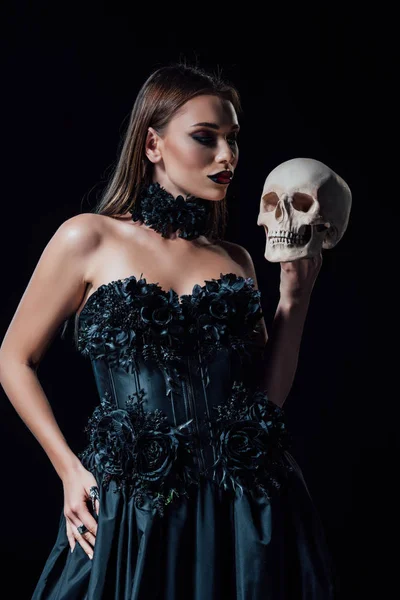 Asustadiza chica vampiro en vestido gótico negro sosteniendo cráneo humano aislado en negro - foto de stock