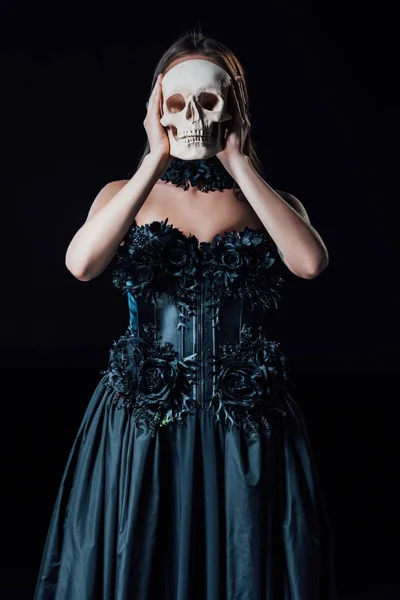 Asustadiza chica vampiro en vestido gótico negro sosteniendo cráneo humano en frente de la cara aislado en negro - foto de stock