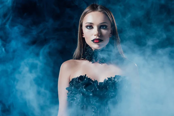 Asustadiza chica vampiro en vestido gótico negro sobre fondo negro con humo - foto de stock