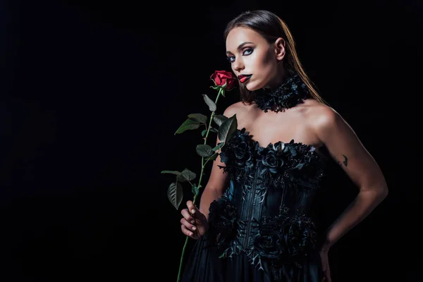 Asustadiza chica vampiro en vestido gótico negro sosteniendo rosa roja aislada en negro - foto de stock