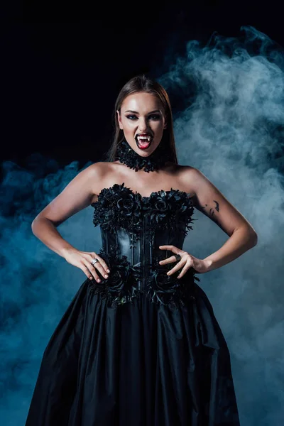 Страшна вампірська дівчина з іклами в чорній готичній сукні на чорному тлі з димом — стокове фото
