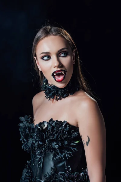 Asustadiza chica vampiro con colmillos en vestido gótico negro con la boca abierta mirando hacia otro lado aislado en negro - foto de stock