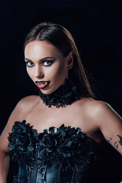 Gruseliges Vampirmädchen mit Reißzähnen in schwarzem Gothikkleid, das isoliert auf schwarz in die Kamera blickt — Stockfoto
