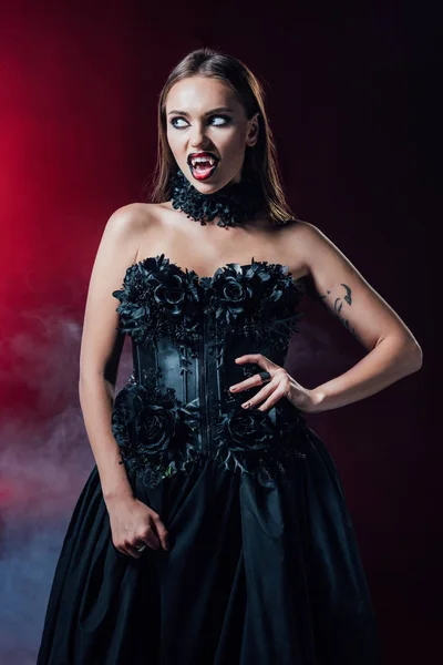 Asustadiza chica vampiro con colmillos en vestido gótico negro sobre fondo negro con humo - foto de stock