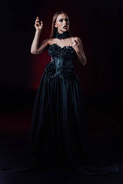 Assustador vampiro menina com presas em preto vestido gótico no fundo preto — Fotografia de Stock
