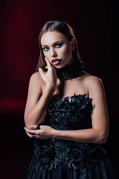 Scary vampire girl with fangs in black gothic dress touching face on black background — Stock Photo