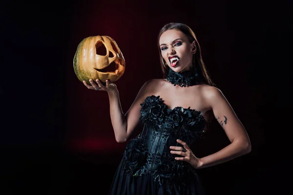 Gruseliges Vampirmädchen mit Reißzähnen in schwarzem Gothikkleid, das einen Halloween-Kürbis auf schwarzem Hintergrund hält — Stockfoto