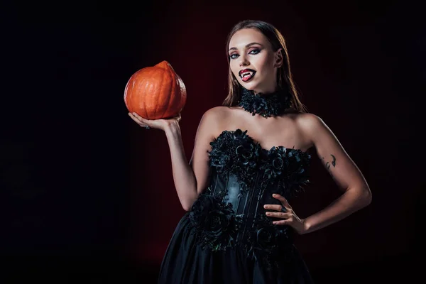 Gruseliges Vampirmädchen mit Reißzähnen in schwarzem Gothikkleid, das einen Halloween-Kürbis auf schwarzem Hintergrund hält — Stockfoto