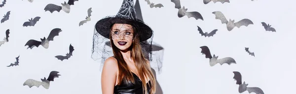 Colpo panoramico di ragazza sorridente in costume strega nera Halloween guardando lontano vicino al muro bianco con pipistrelli decorativi — Foto stock