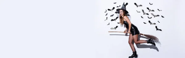 Plan panoramique de fille en costume d'Halloween sorcière noire sur balai près du mur blanc avec des chauves-souris décoratives — Photo de stock