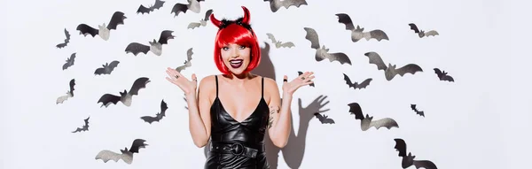 Panoramaaufnahme eines aufgeregten Mädchens im Teufel-Halloween-Kostüm nahe weißer Wand mit dekorativen Fledermäusen — Stockfoto