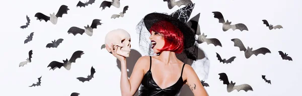 Panoramic shot of girl in black witch Halloween costume with red hair holding skull near white wall with decorative bats — Stock Photo