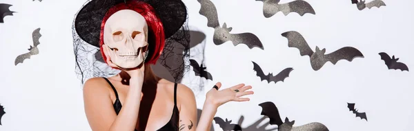 Plano panorámico de chica en traje de Halloween bruja negro con pelo rojo celebración de cráneo en frente de la cara cerca de la pared blanca con murciélagos decorativos - foto de stock