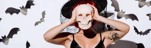Plano panorámico de chica en traje de Halloween bruja negro con pelo rojo celebración de cráneo en frente de la cara cerca de la pared blanca con murciélagos decorativos - foto de stock
