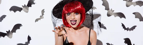 Plano panorámico de chica en traje de Halloween bruja negro con pelo rojo cerca de la pared blanca con murciélagos decorativos - foto de stock