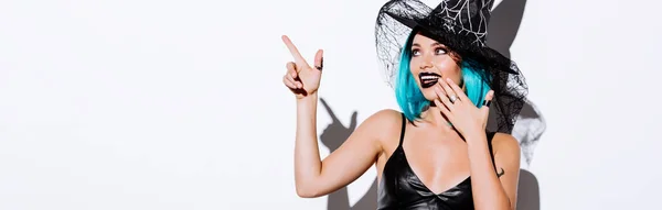 Plano panorámico de chica sonriente en traje de Halloween bruja negro con el pelo azul señalando con el dedo sobre fondo blanco - foto de stock