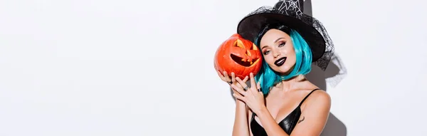 Plano panorámico de chica sonriente en traje de Halloween bruja negro con pelo azul celebración espeluznante calabaza tallada sobre fondo blanco - foto de stock