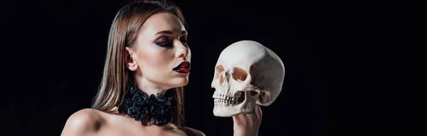 Panoramische Aufnahme eines beängstigenden Vampirmädchens in schwarzem Gothic-Kleid, das den menschlichen Schädel isoliert auf schwarz betrachtet — Stockfoto