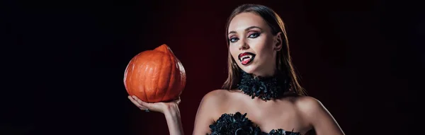 Panoramic shot of scary vampire girl with fangs in black gothic dress holding Halloween pumpkin on black background — Stock Photo