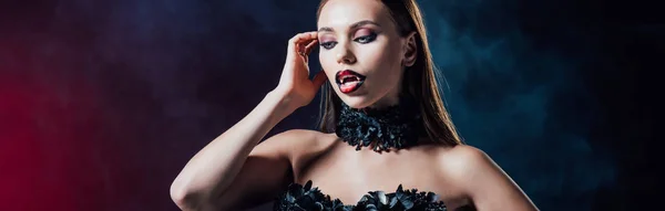 Panoramic shot of scary vampire girl with fangs in black gothic dress on black background with smoke — Stock Photo