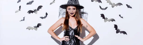 Panoramic shot of girl in black witch Halloween costume with hands on hips near white wall with decorative bats — Stock Photo