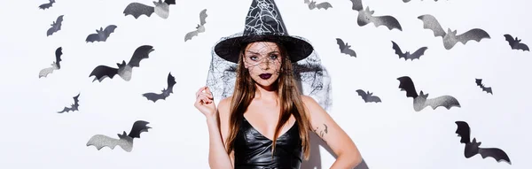 Plano panorámico de chica en traje de Halloween bruja negro cerca de la pared blanca con murciélagos decorativos - foto de stock
