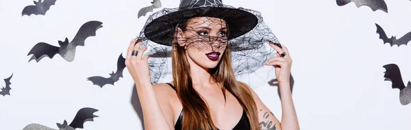 Colpo panoramico di ragazza in costume strega nera Halloween tenuta velo sul cappello vicino muro bianco con pipistrelli decorativi — Foto stock