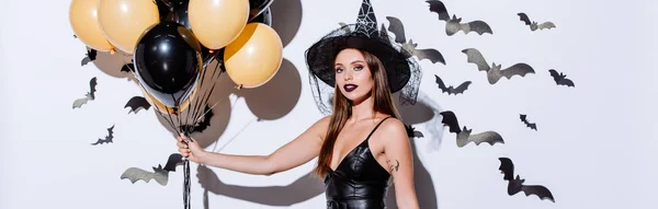 Панорамный снимок девушки в костюме чёрной ведьмы на Хэллоуин, держащей воздушные шары возле белой стены с декоративными летучими мышами — стоковое фото