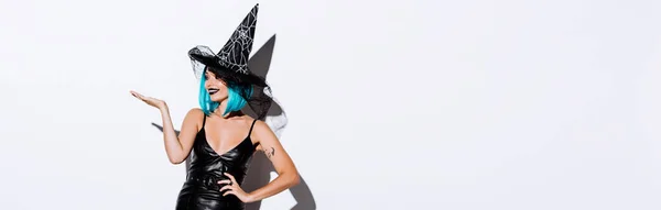 Plano panorámico de chica feliz en traje de Halloween bruja negro con el pelo azul señalando con la mano sobre fondo blanco - foto de stock