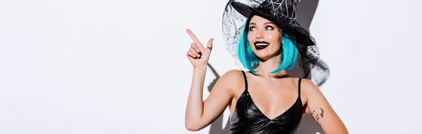 Colpo panoramico di ragazza sorridente in costume nero strega Halloween con i capelli blu puntando con il dito su sfondo bianco — Foto stock