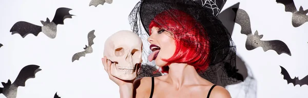 Panoramic shot of girl in black witch Halloween costume with red hair licking skull near white wall with decorative bats — Stock Photo
