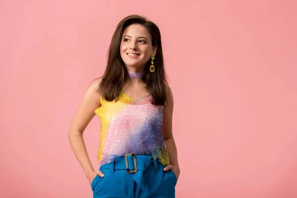 Sonriente joven y elegante chica disco con las manos en los bolsillos mirando hacia otro lado aislado en rosa - foto de stock