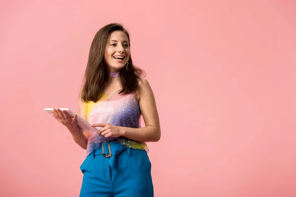 Aufgeregtes junges stilvolles Disco-Mädchen mit digitalem Tablet, das isoliert auf rosa wegschaut — Stockfoto